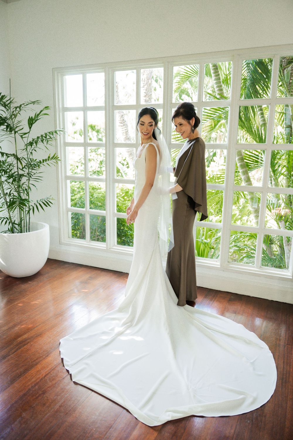 Modern sunlit room with white wedding gown trailing elegantly across dark hardwood floors next to tropical window views.