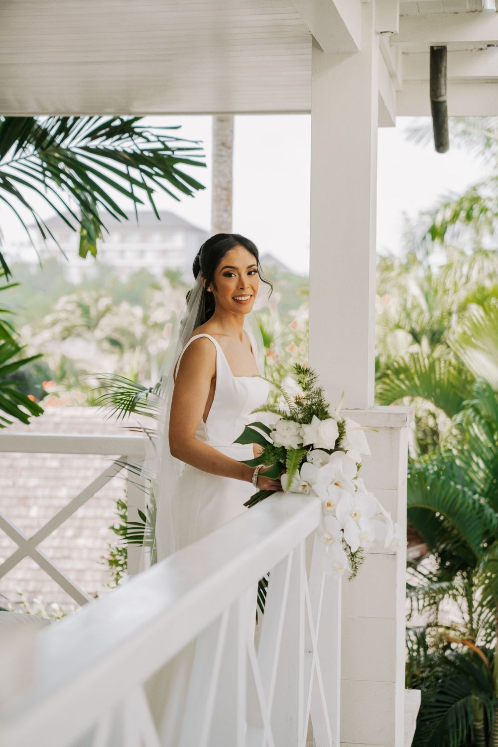 A bride in a white strapless gown poses on a porch with tropical plants, holding a bouquet of white orchids.
