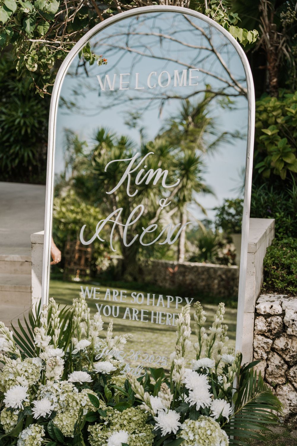 Elegant wedding welcome signs with white floral decorations and mirrored surface displayed in an outdoor garden setting.