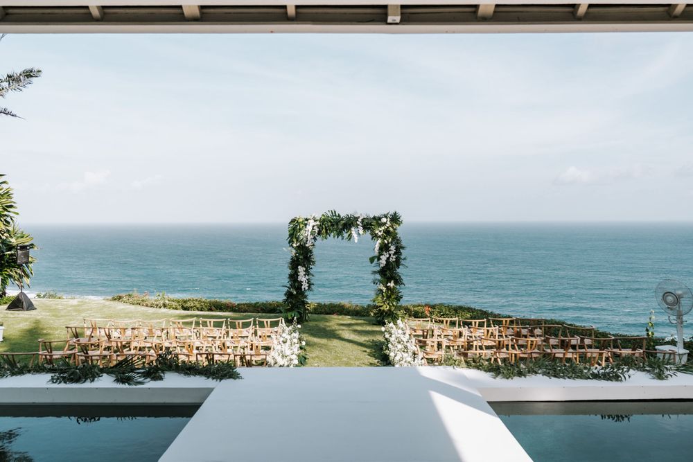 Ocean view wedding ceremony setup with floral arch and white chairs on clifftop overlooking the sea.