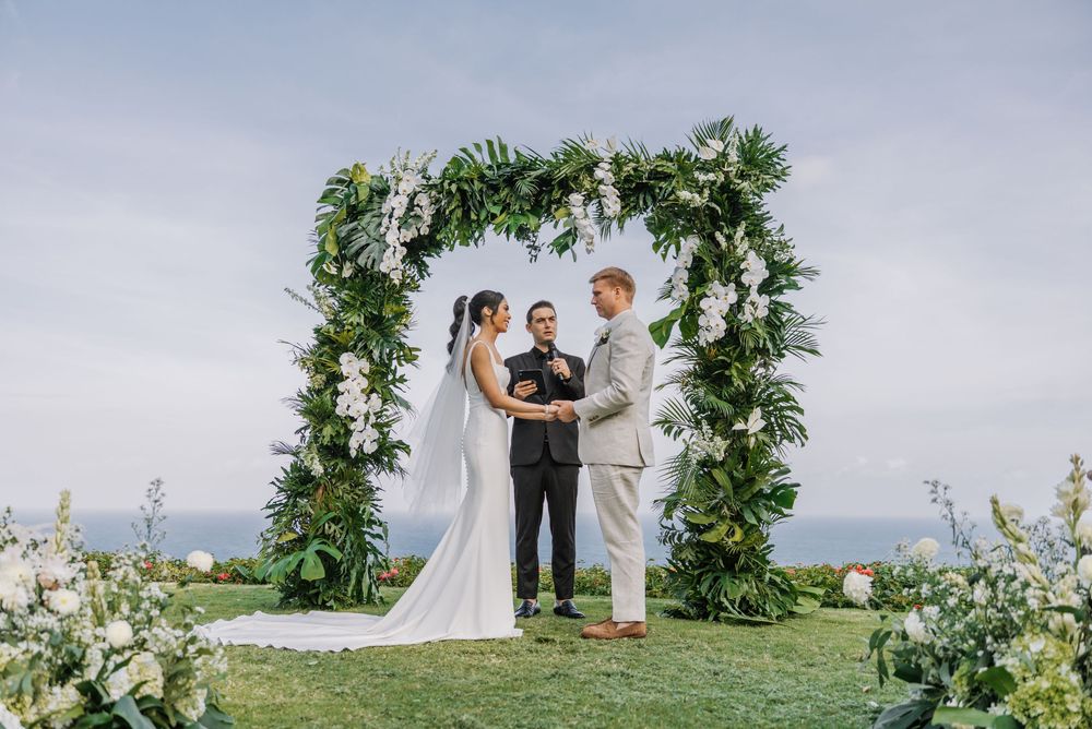 Outdoor wedding ceremony takes place under a lush green floral arch with ocean views in the background.