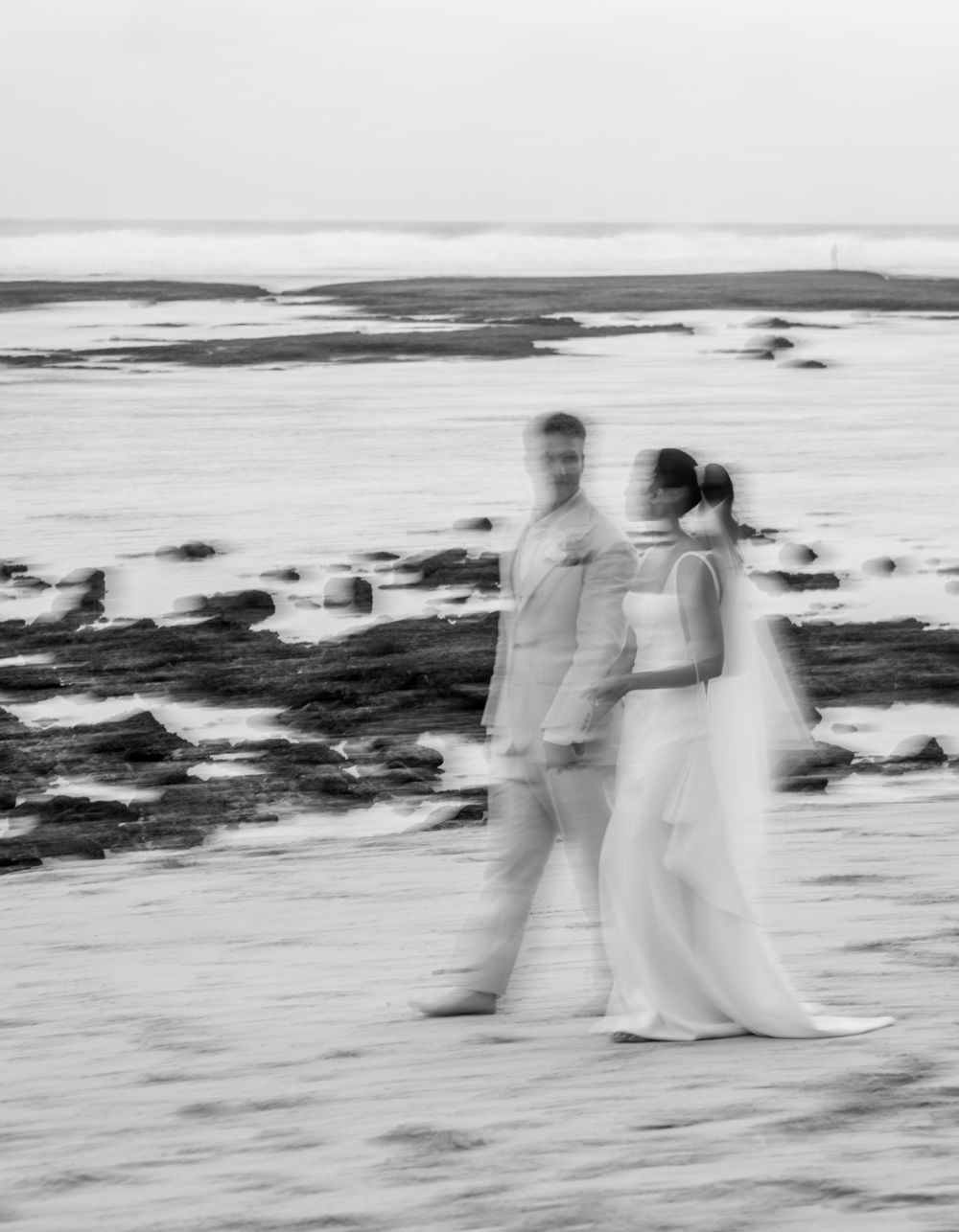 Blurred motion black and white photograph of figures walking along a misty beach shoreline at sunset.
