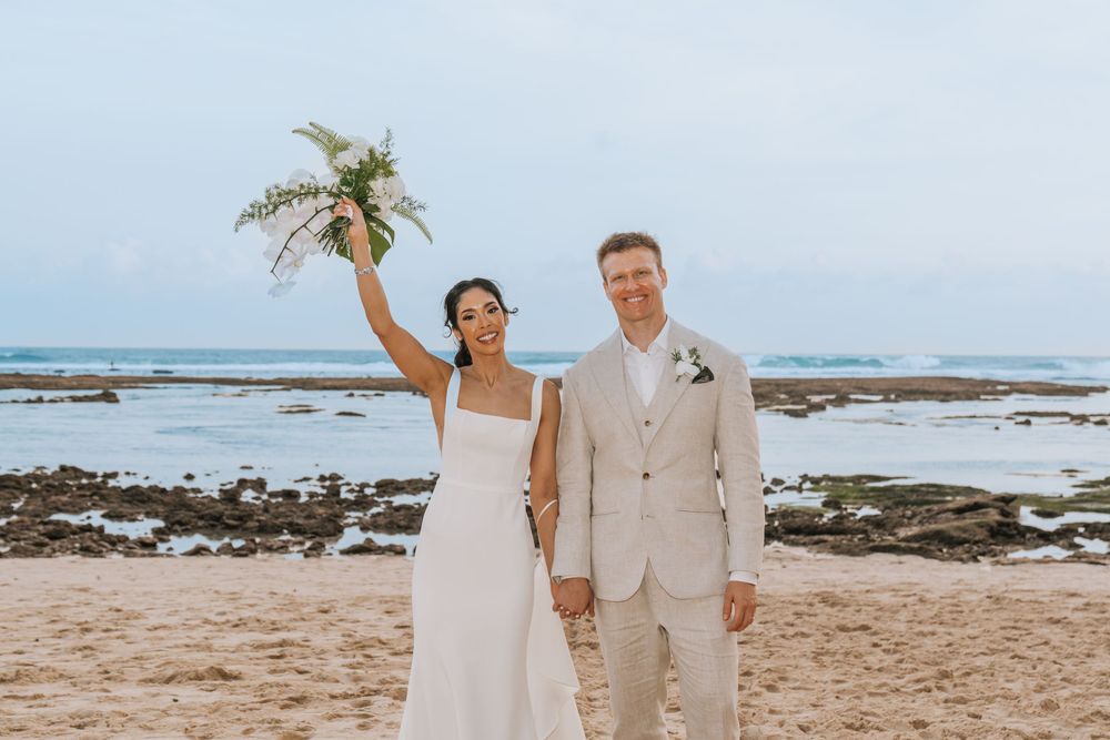 A beachside wedding ceremony with a couple embracing on a rocky Hawaiian shoreline with ocean waves in the background.