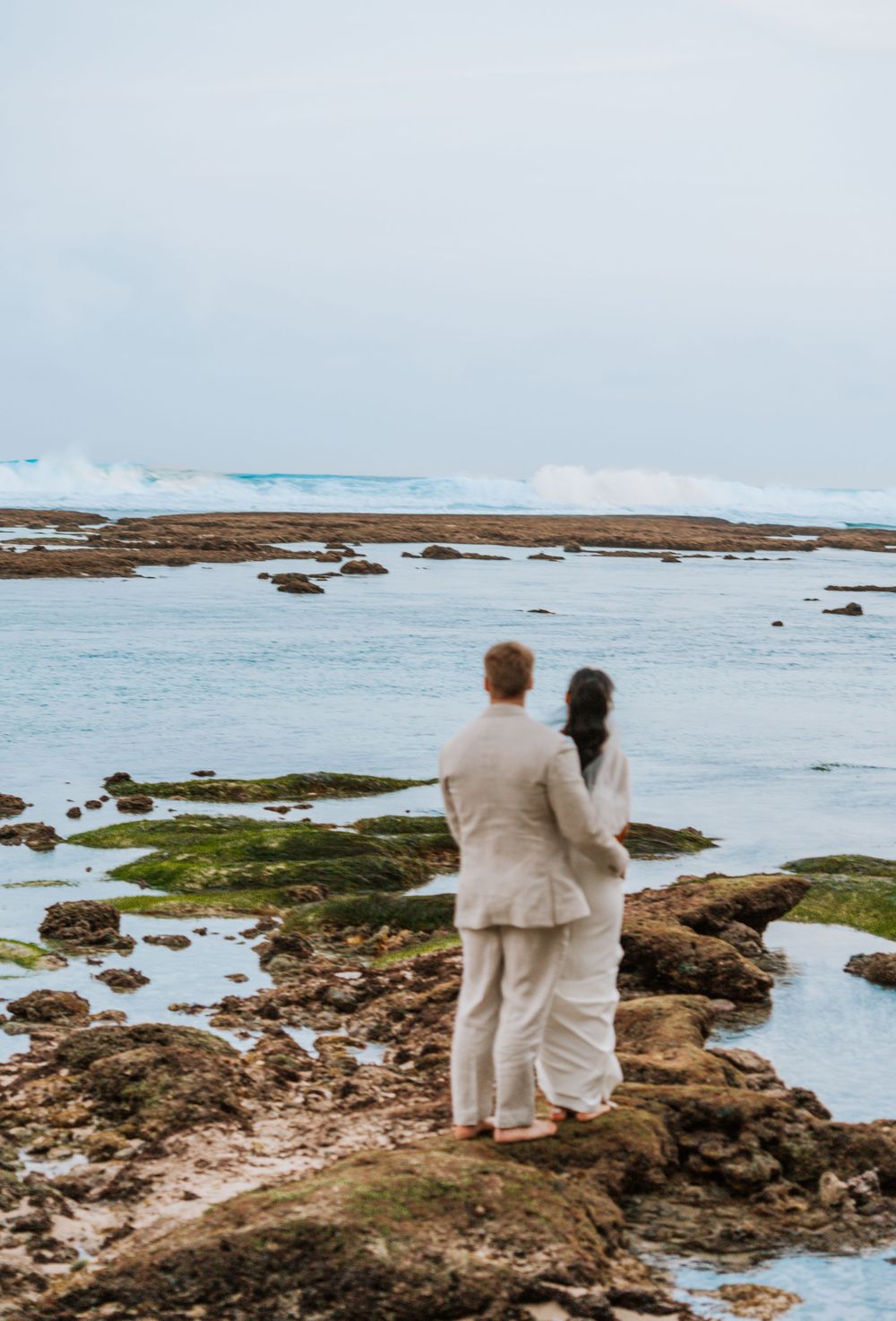 Two people in light-colored clothing stand together on rocky coastal shoreline overlooking blue ocean waters and mossy rocks.