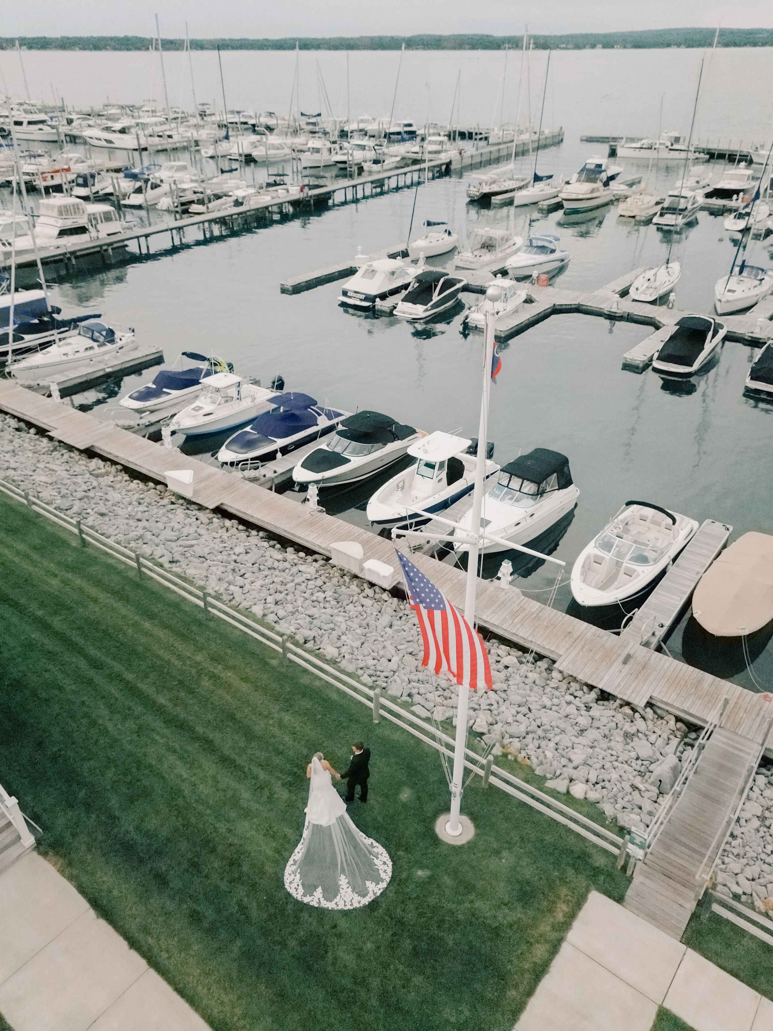 Marina view with boats docked along stone walkways, green lawn, and American flags visible in the foreground.