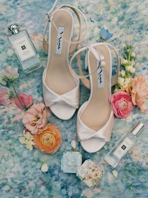 Elegant white bridal shoes and perfume bottles arranged with pink and peach flowers on a blue marbled surface.