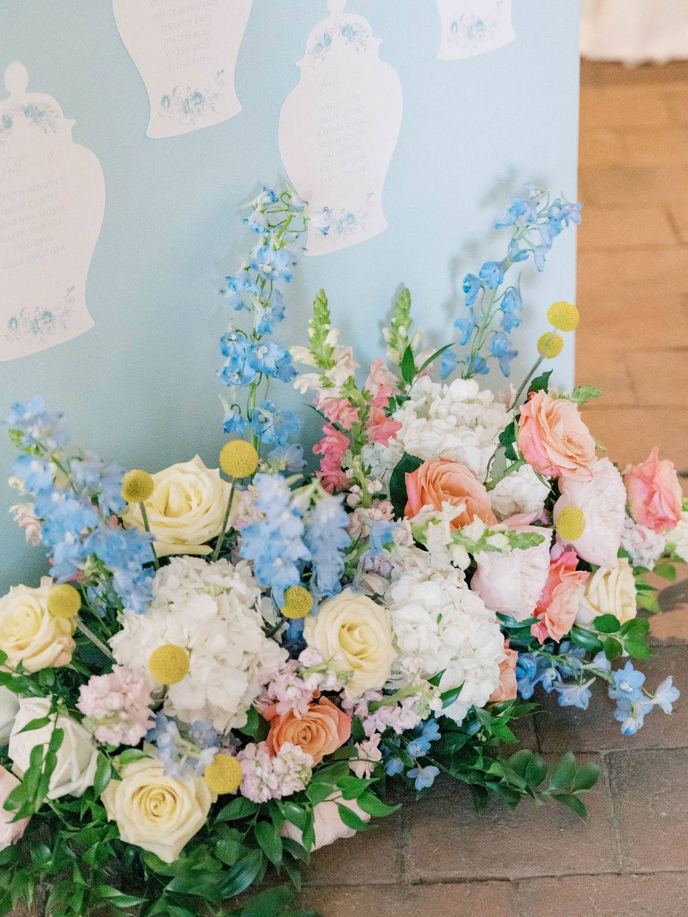 Elegant floral arrangement featuring pink and white roses, delphiniums, and mixed flowers against a light blue backdrop.