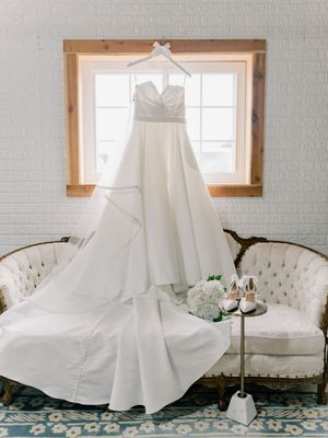 White wedding dress hanging elegantly against white brick wall with natural window light.