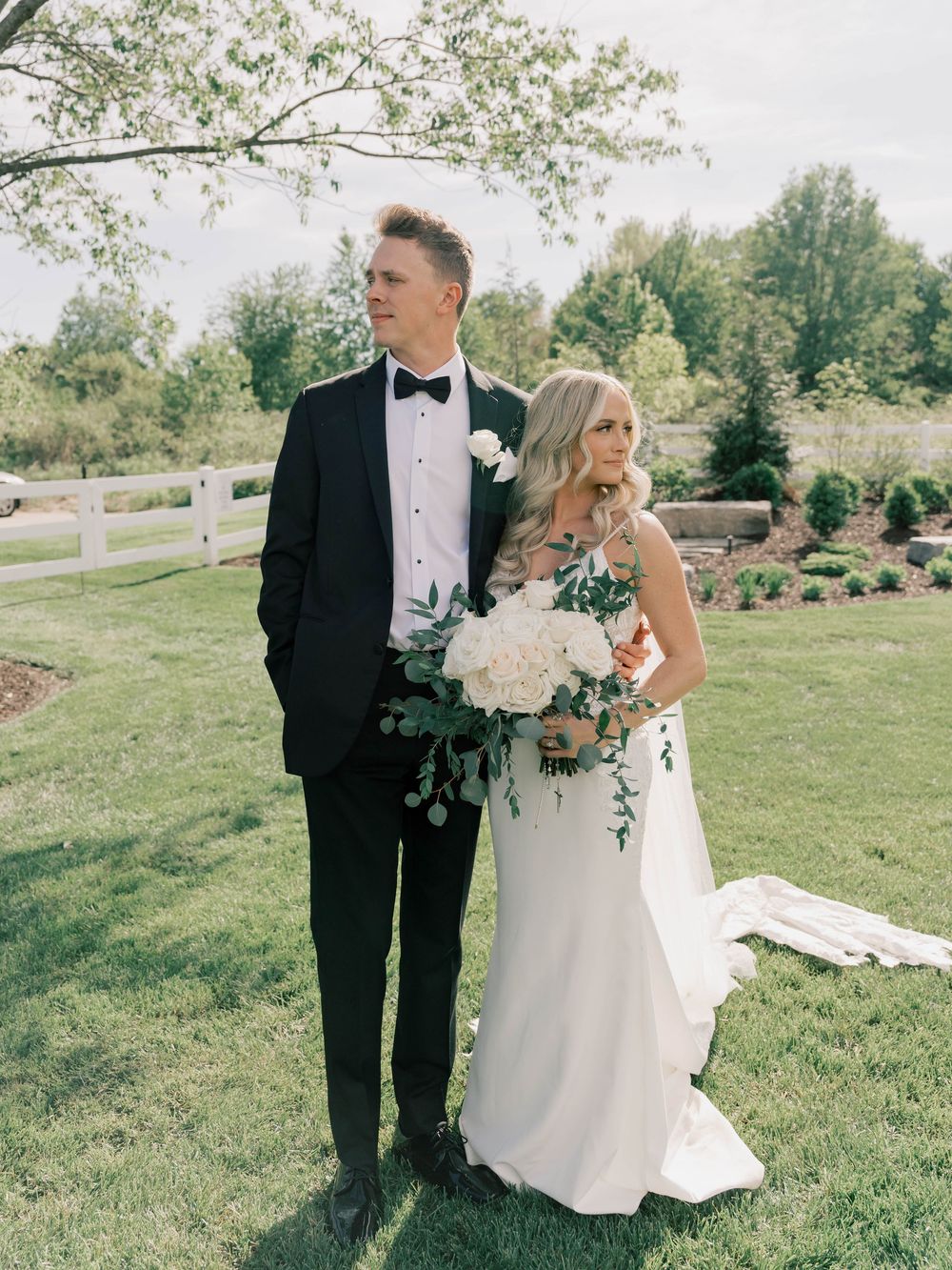 Elegant wedding couple poses in a garden setting with white floral bouquet and classic attire.