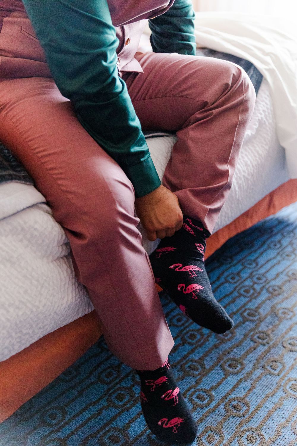 A person wearing green sweater and burgundy pants sits on a patterned carpet wearing fun flamingo socks.