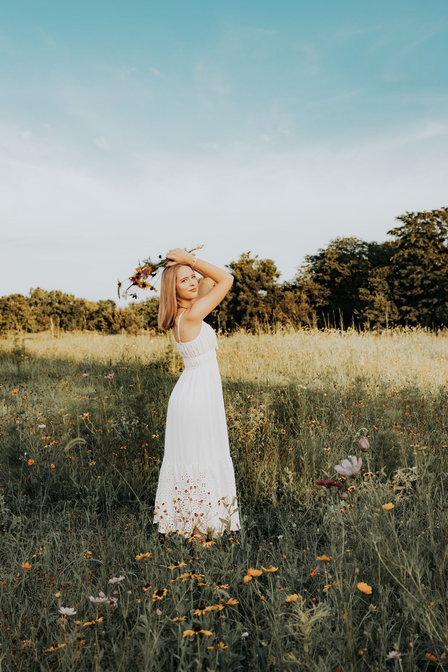 A serene outdoor portrait of someone in a flowing white dress standing in a wildflower meadow during golden hour.