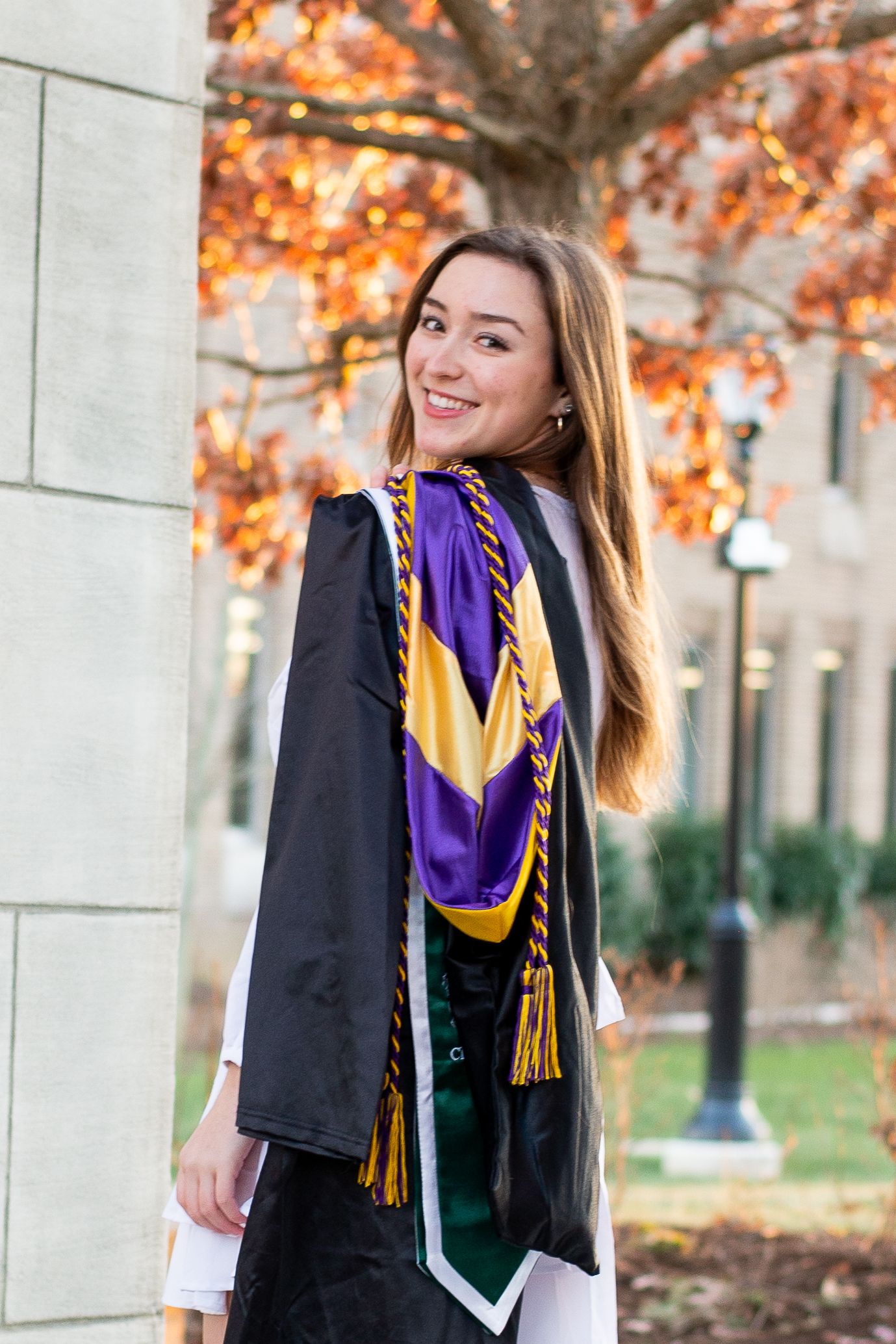 Graduate in black cap and gown stands on path in front of stone church building with green trees on campus.