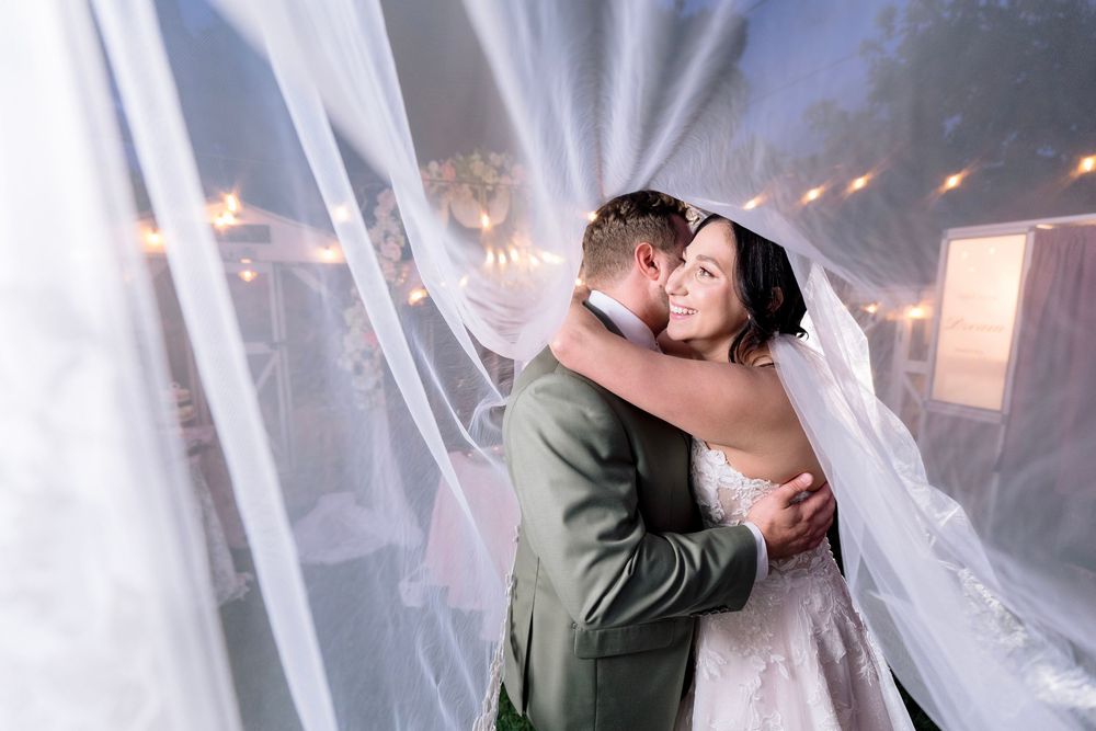 Bride and groom share intimate embrace wrapped in flowing veil under string lights at evening reception.