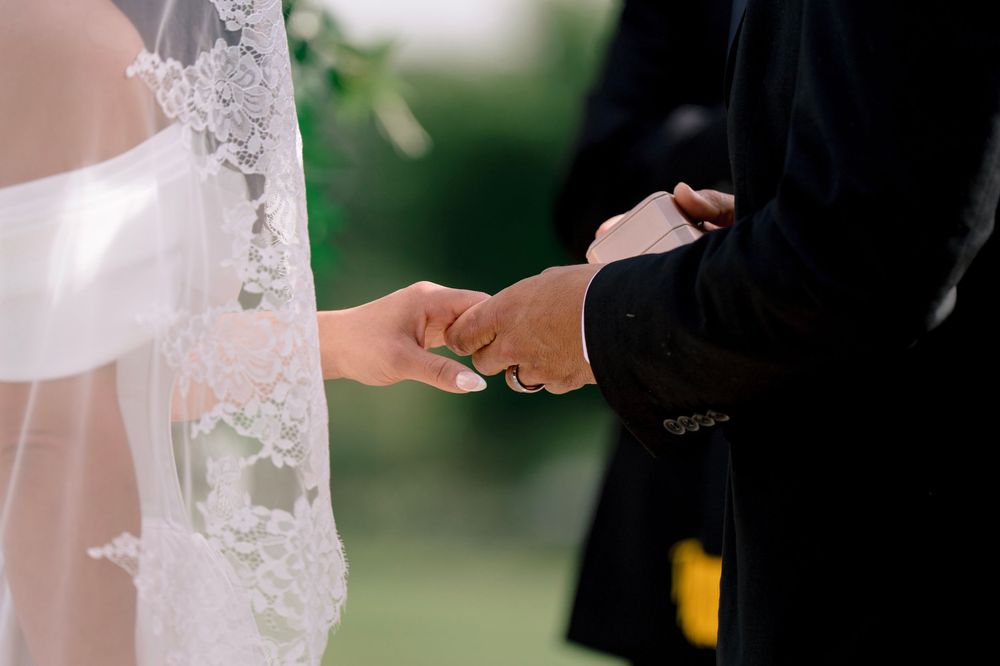 Bride and groom hold hands tenderly during wedding ceremony, bride wearing lace veil and white gown.