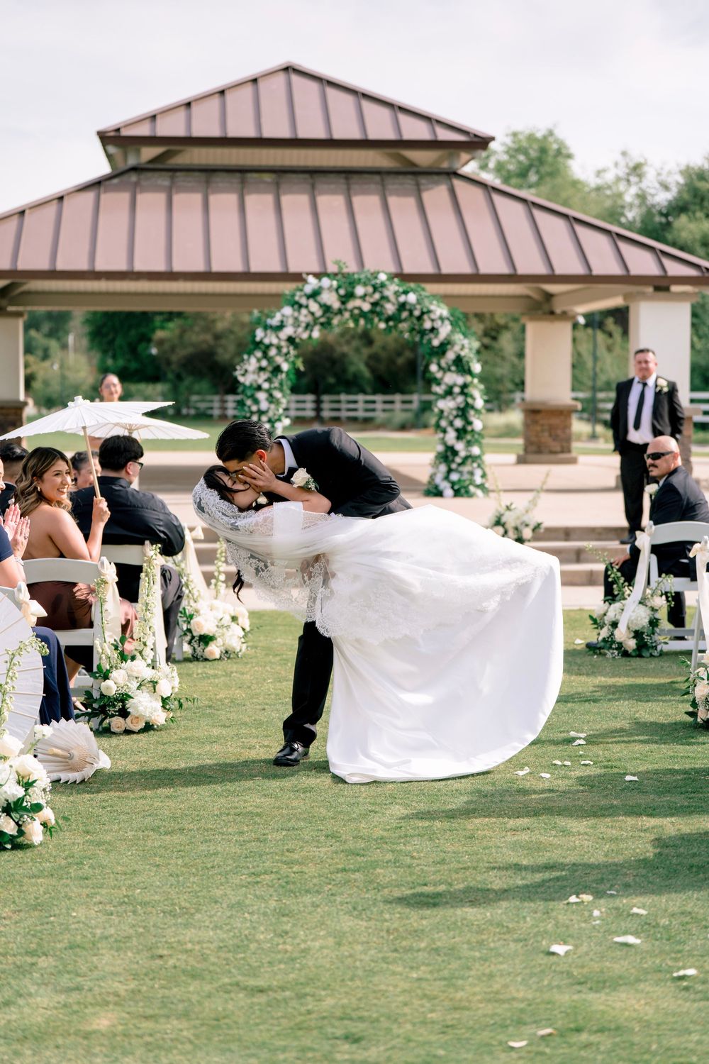 Groom dips bride for romantic kiss in wedding aisle with floral arch and seated guests behind them.