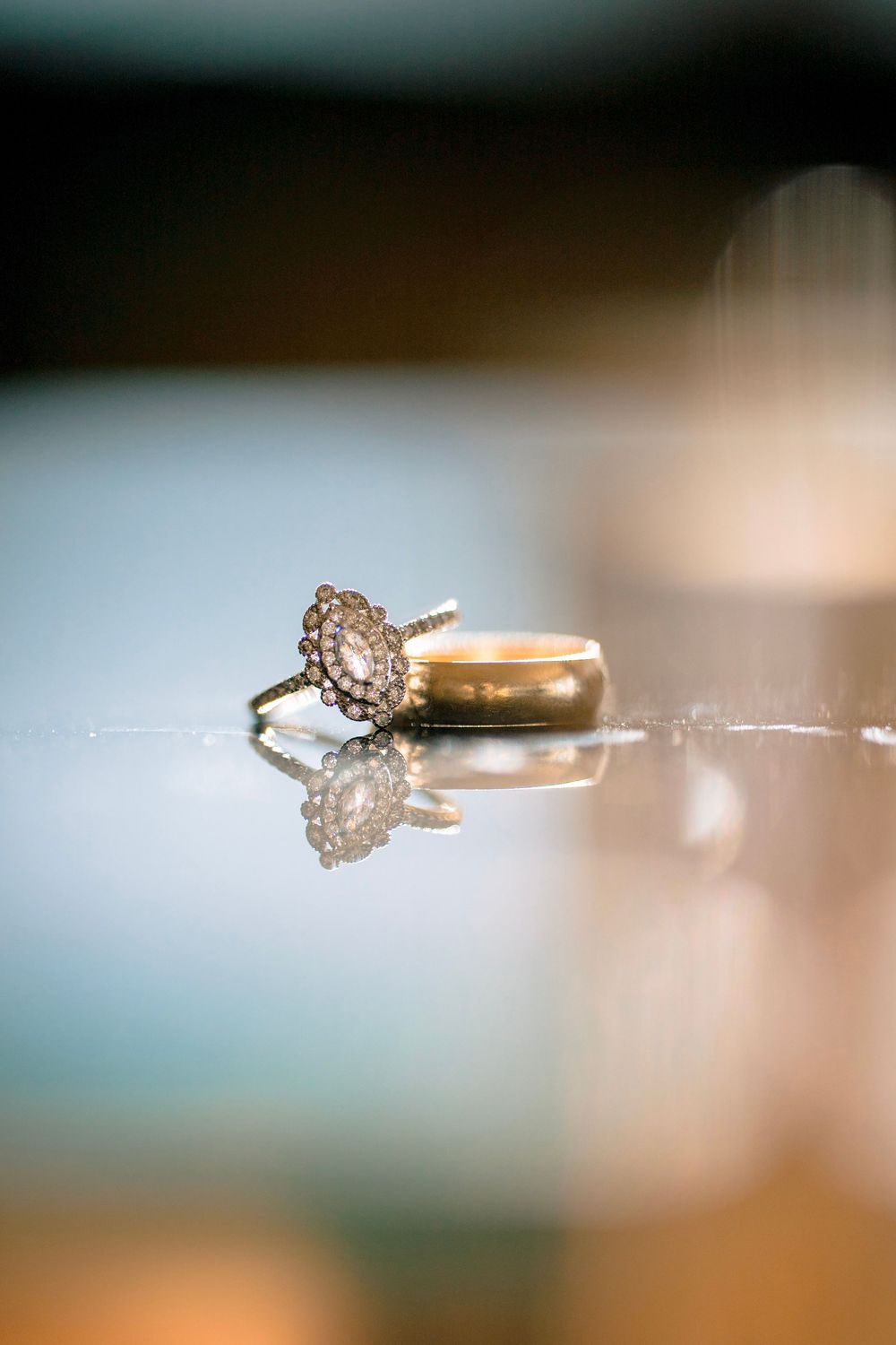 Vintage oval diamond engagement ring with halo beside a gold wedding band on reflective surface.