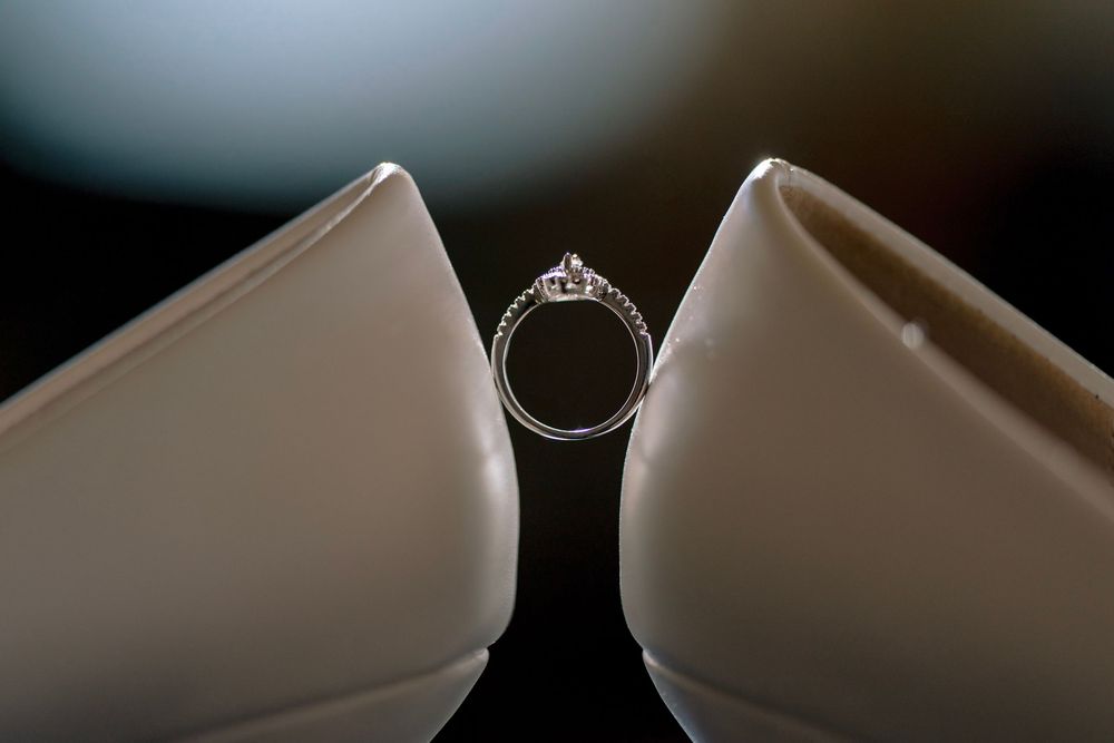 Diamond solitaire engagement ring displayed between two white bridal heels on dark background.