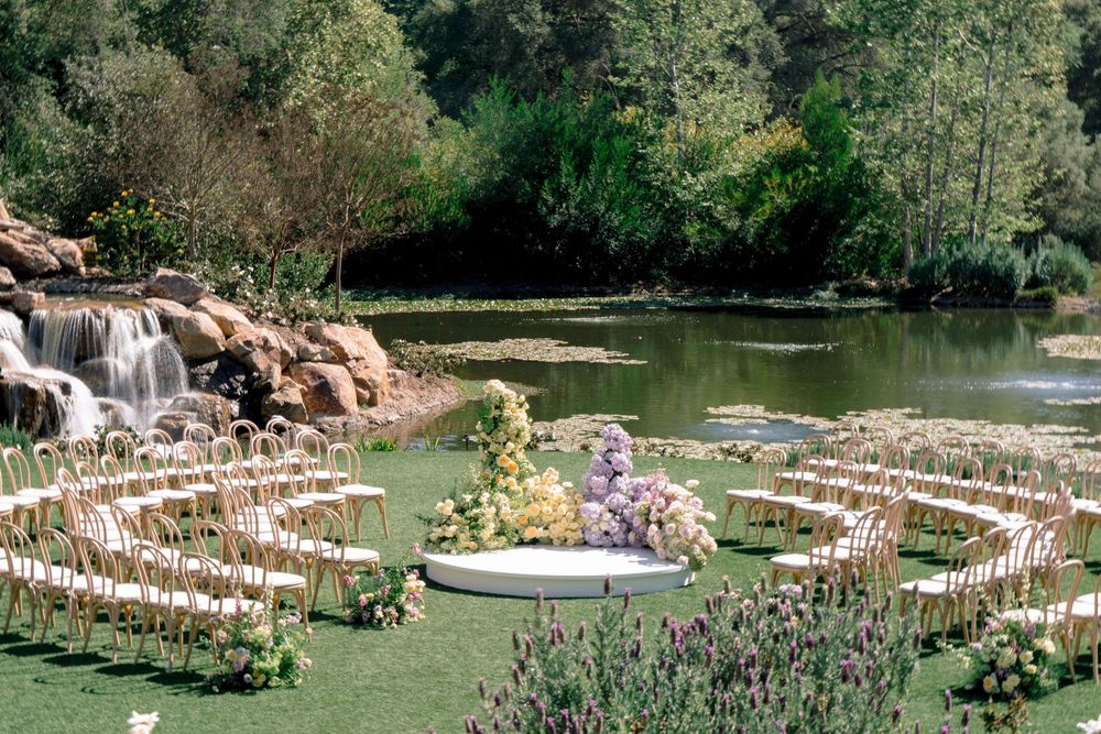 Outdoor wedding ceremony setup with circular seating arrangement, floral centerpiece, waterfall, and lush greenery by a serene pond.
