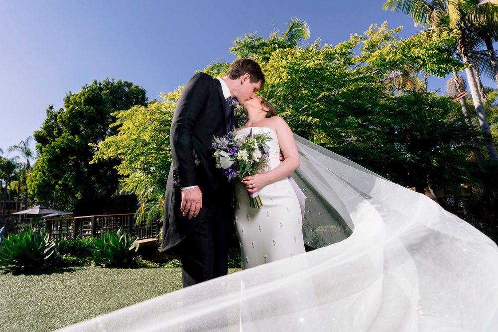 Bride and groom sharing a romantic kiss outdoors with flowing veil and lush green trees behind them.
