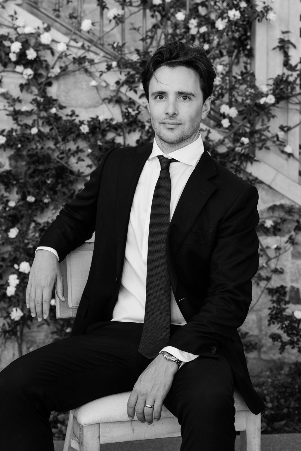 Elegant groom in black suit and tie seated on chair with flowering vines in black and white portrait.