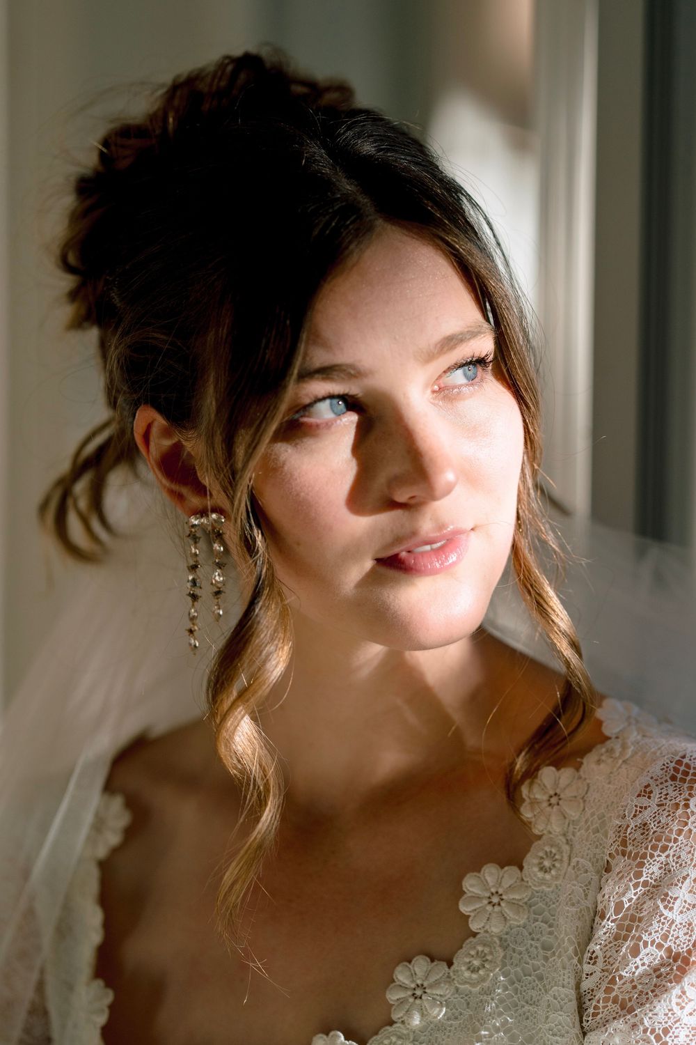 Beautiful bride with updo hairstyle and dangling earrings gazing in natural window light.