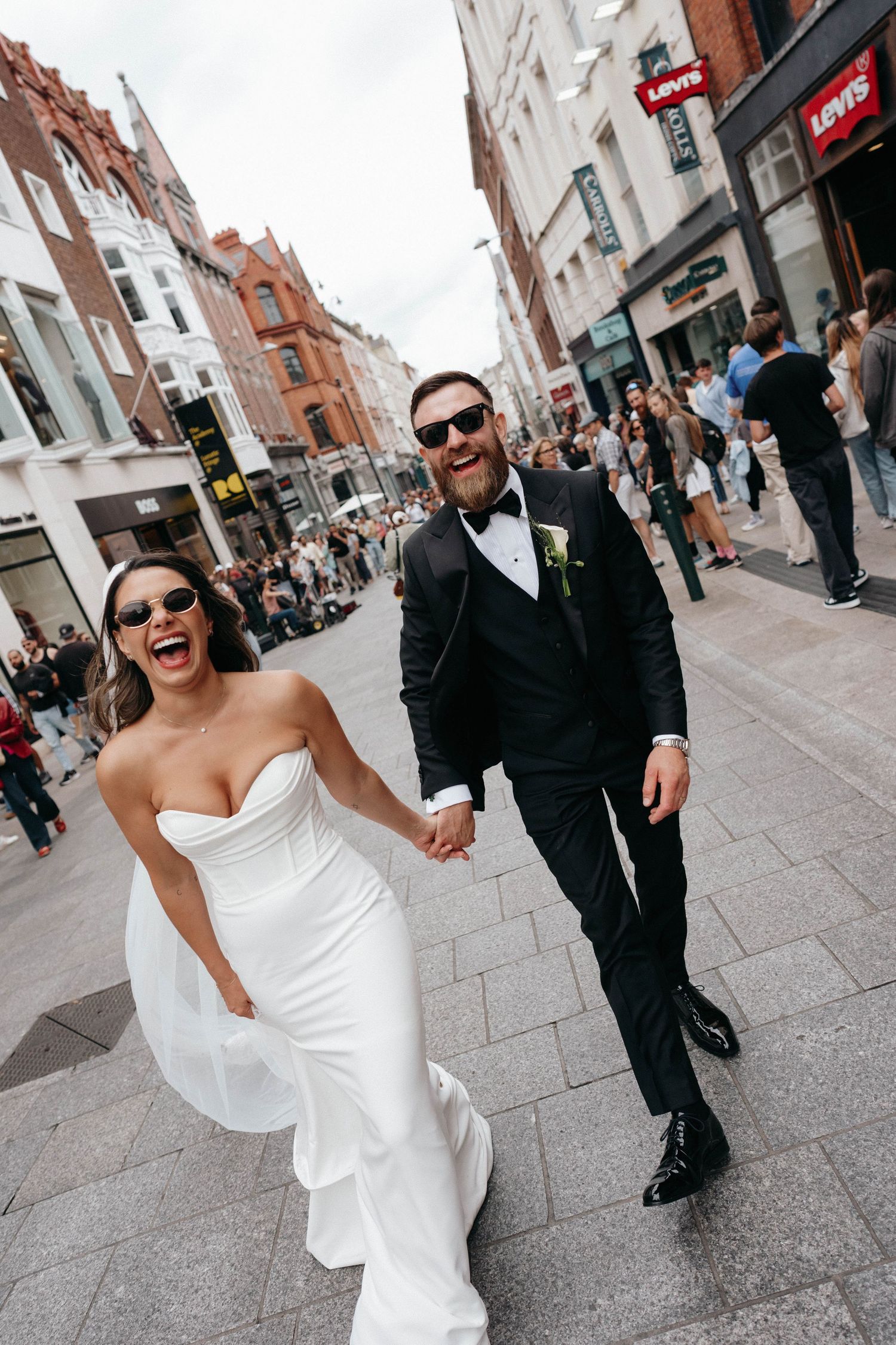 Newlywed couple laughing and walking down a cobblestone street with historic buildings visible in background.