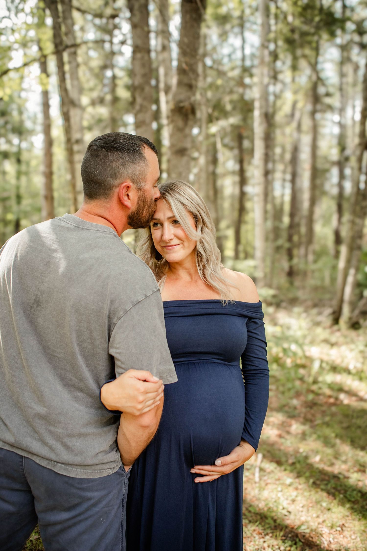 Couple in navy and gray clothing embrace in a sunlit forest setting.