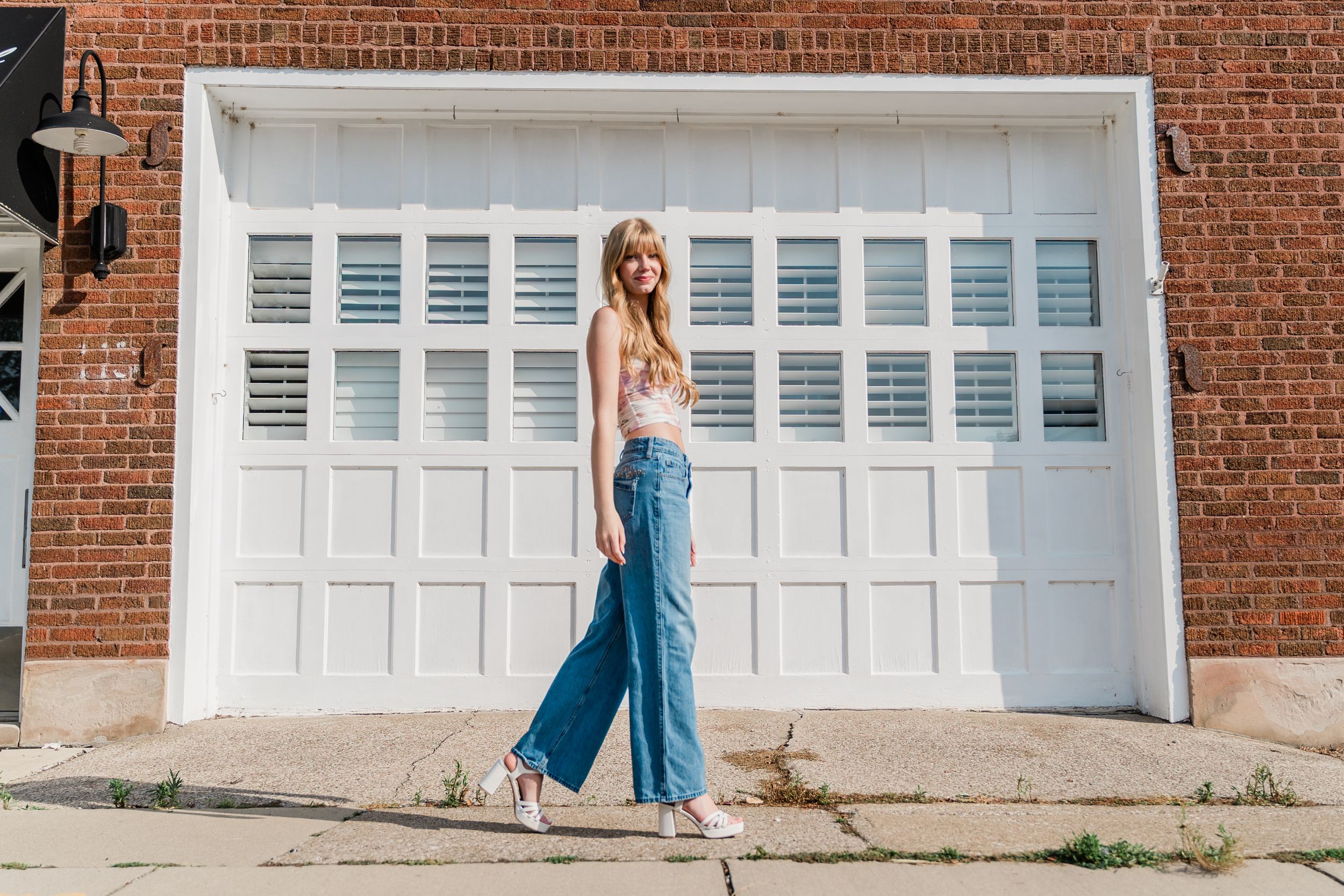 A person wearing a white top and blue wide-leg pants poses in front of a white garage door against a brick wall.