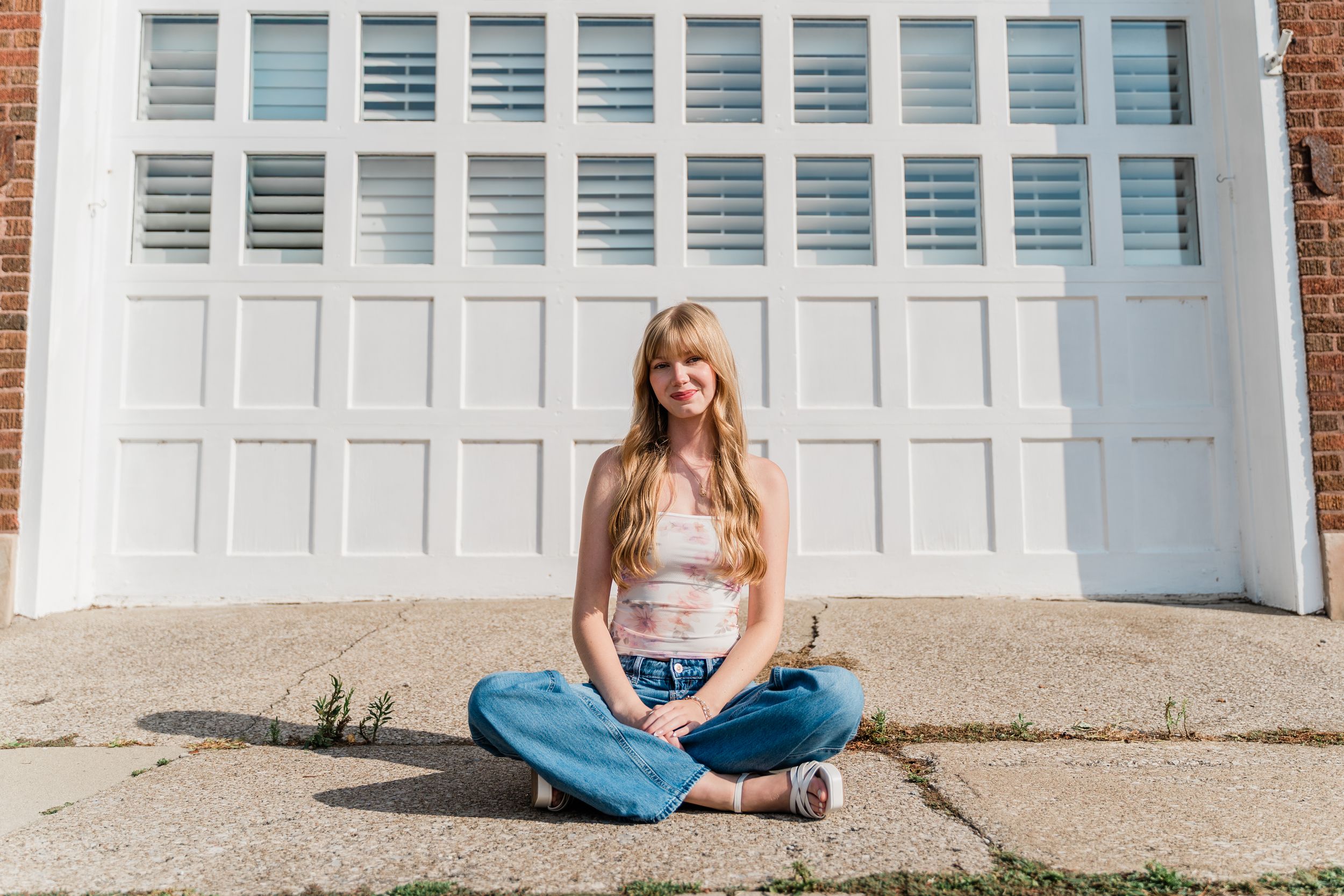 Carefree person in blue jeans and white top sits cross-legged in front of white garage door on sunny day.