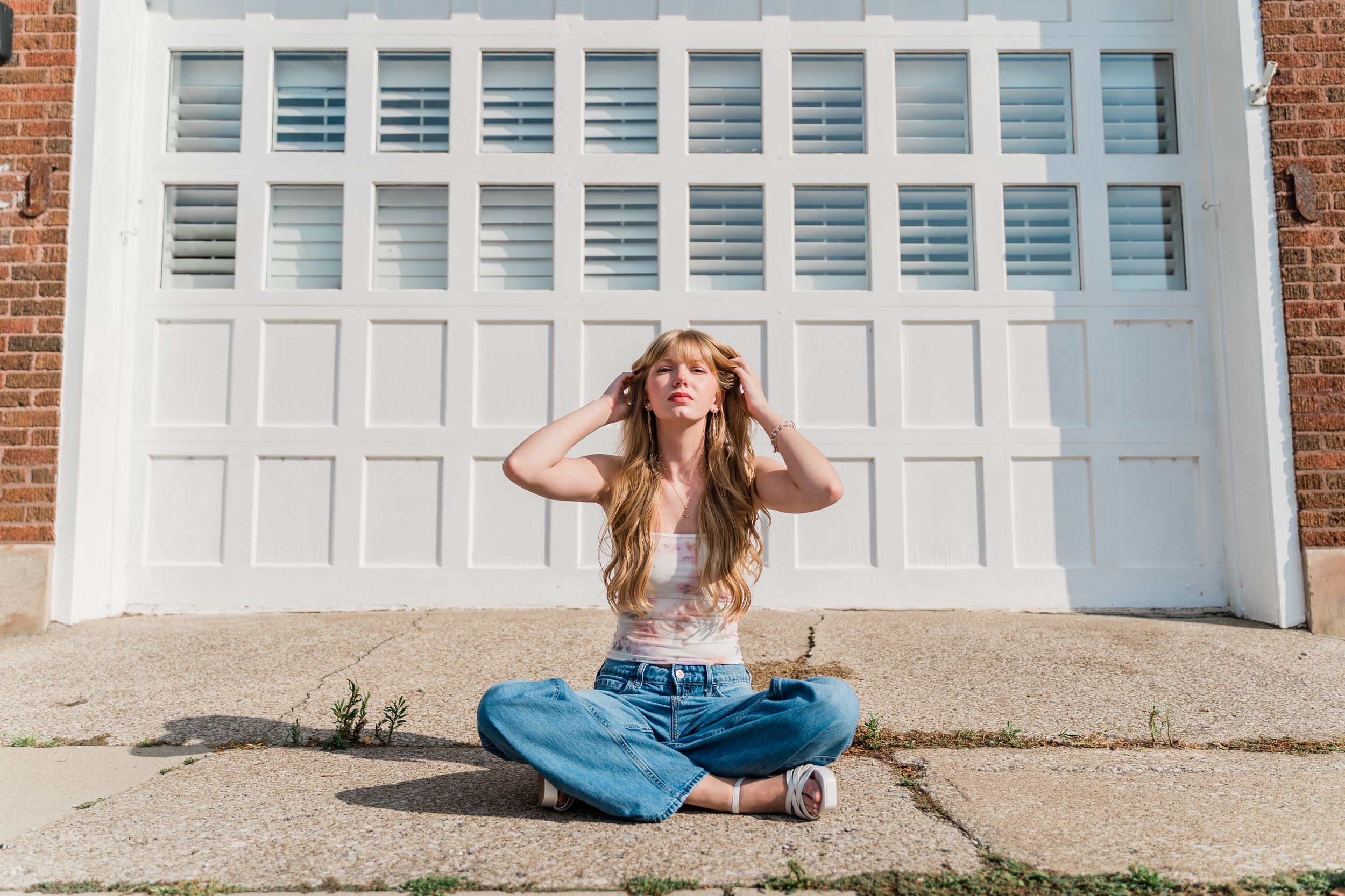 Casual figure in denim and crop top poses playfully while sitting against bright white garage door.