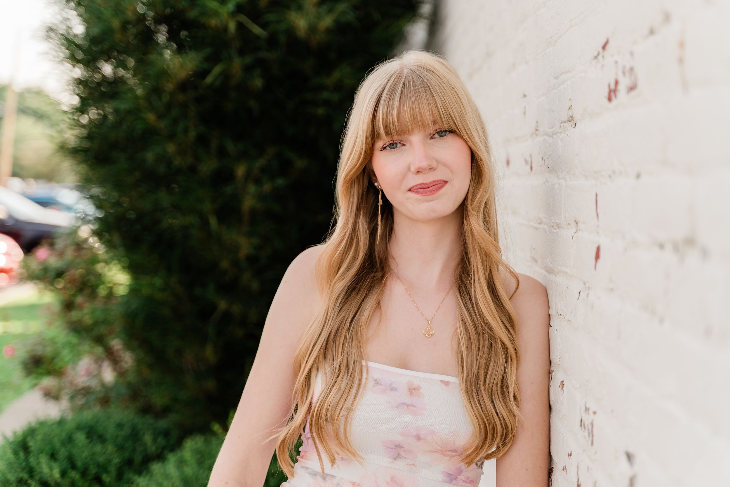 A person with long blonde hair wearing a pink floral dress poses against a white brick wall outdoors.