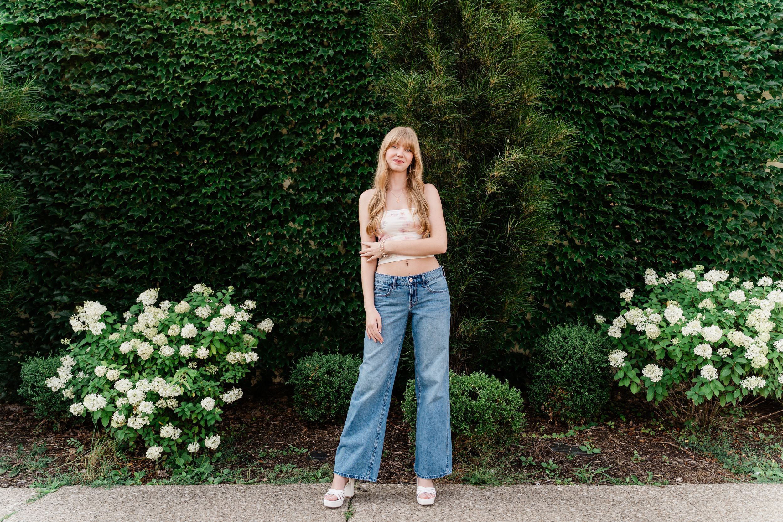 A casual summer fashion photo of someone in a white top and relaxed jeans standing against a green hedge with flowers.