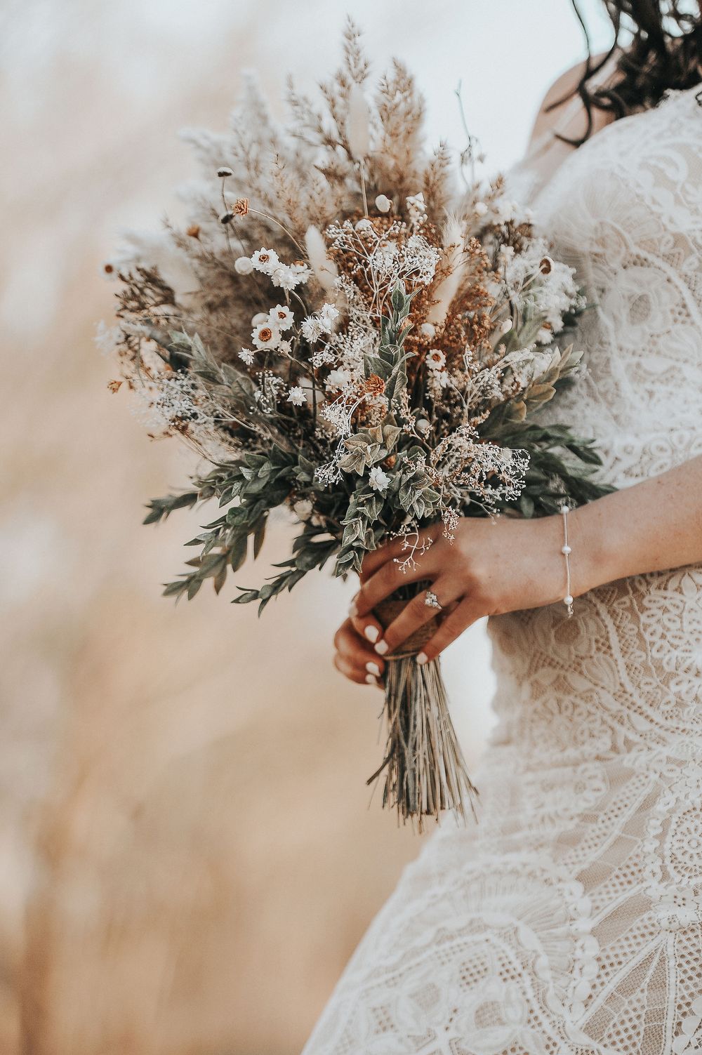 Rustic wedding bouquet featuring dried flowers, greenery and textured elements in neutral tones.
