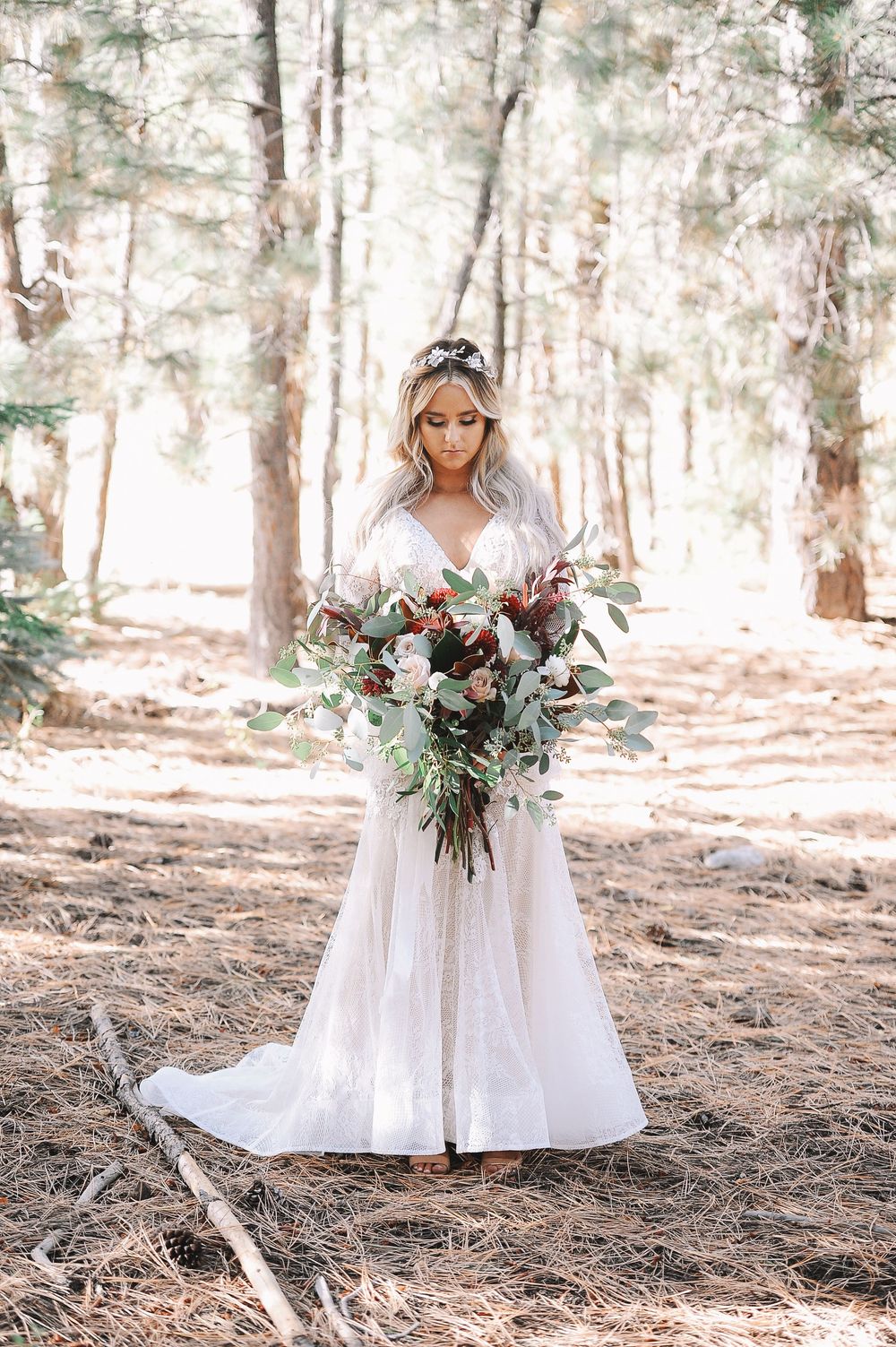 Bride in a flowing white dress holds a bouquet while posing in a sunlit pine forest setting.