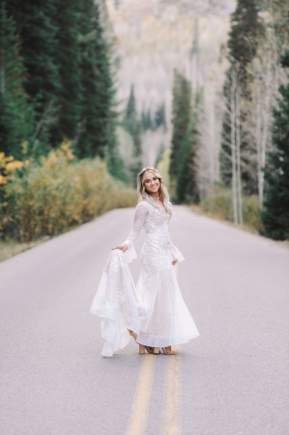Woman in white dress twirls on a mountain road surrounded by evergreen trees and autumn foliage.