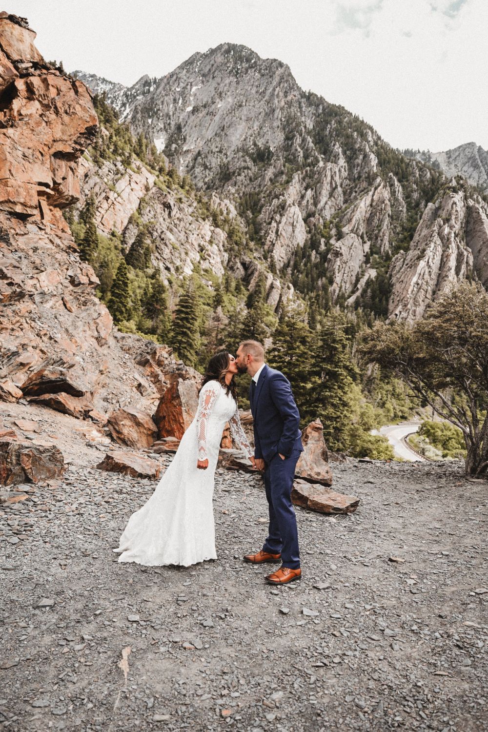 Couple shares an intimate moment on a rocky mountain ledge with dramatic peaks in the background.