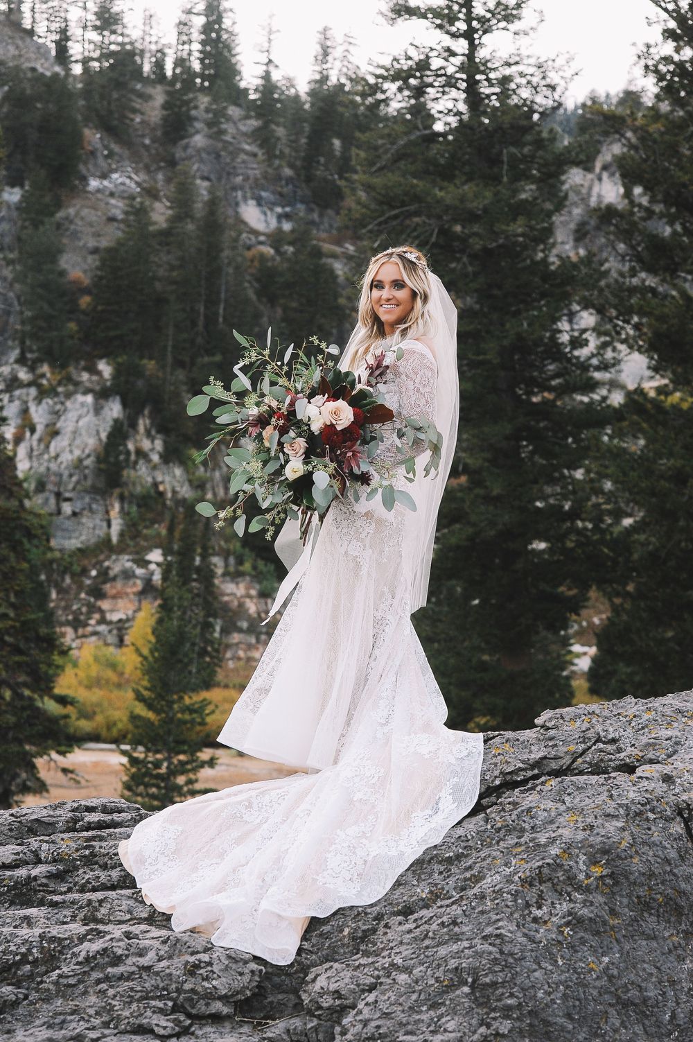 Bride in lace dress holds a burgundy bouquet while standing on rocky mountain terrain.