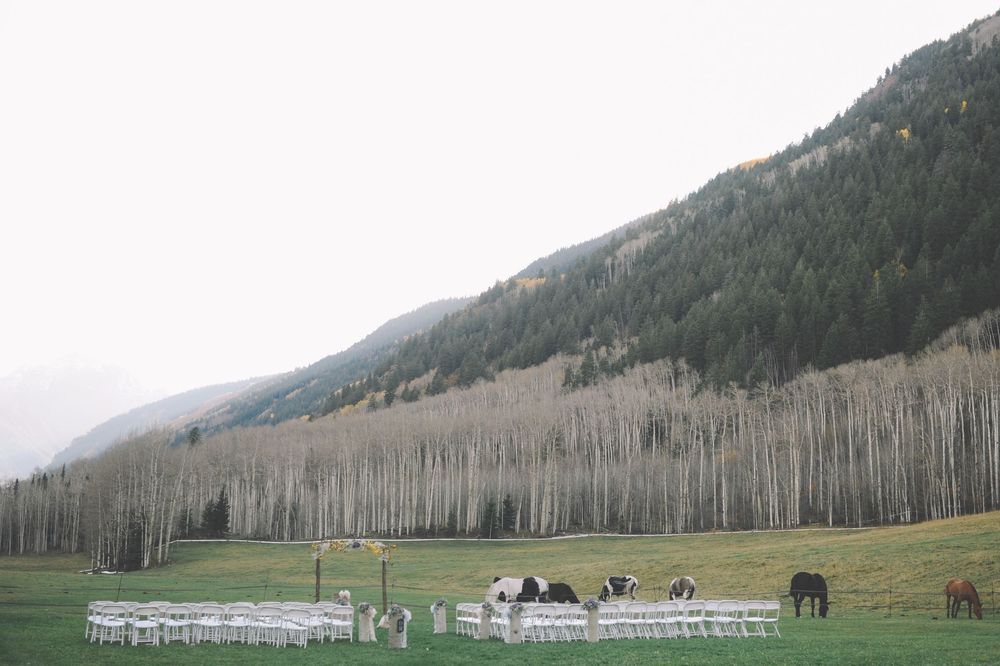 Mountain wedding venue with white chairs set up in meadow and grazing horses in distance.