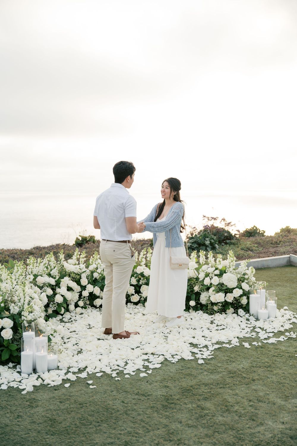 Couple in white outfits stands by elegant floral decor on grassy cliffside overlooking ocean at sunset.