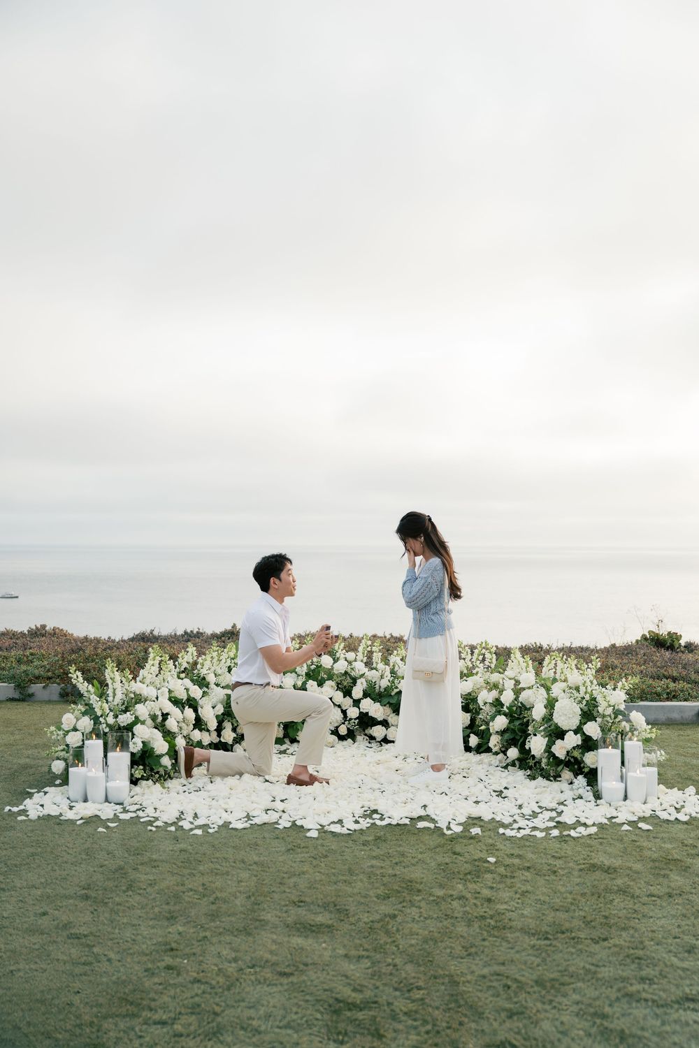 A couple stands by a stunning white floral installation on a grassy hilltop overlooking an ocean view at sunset.