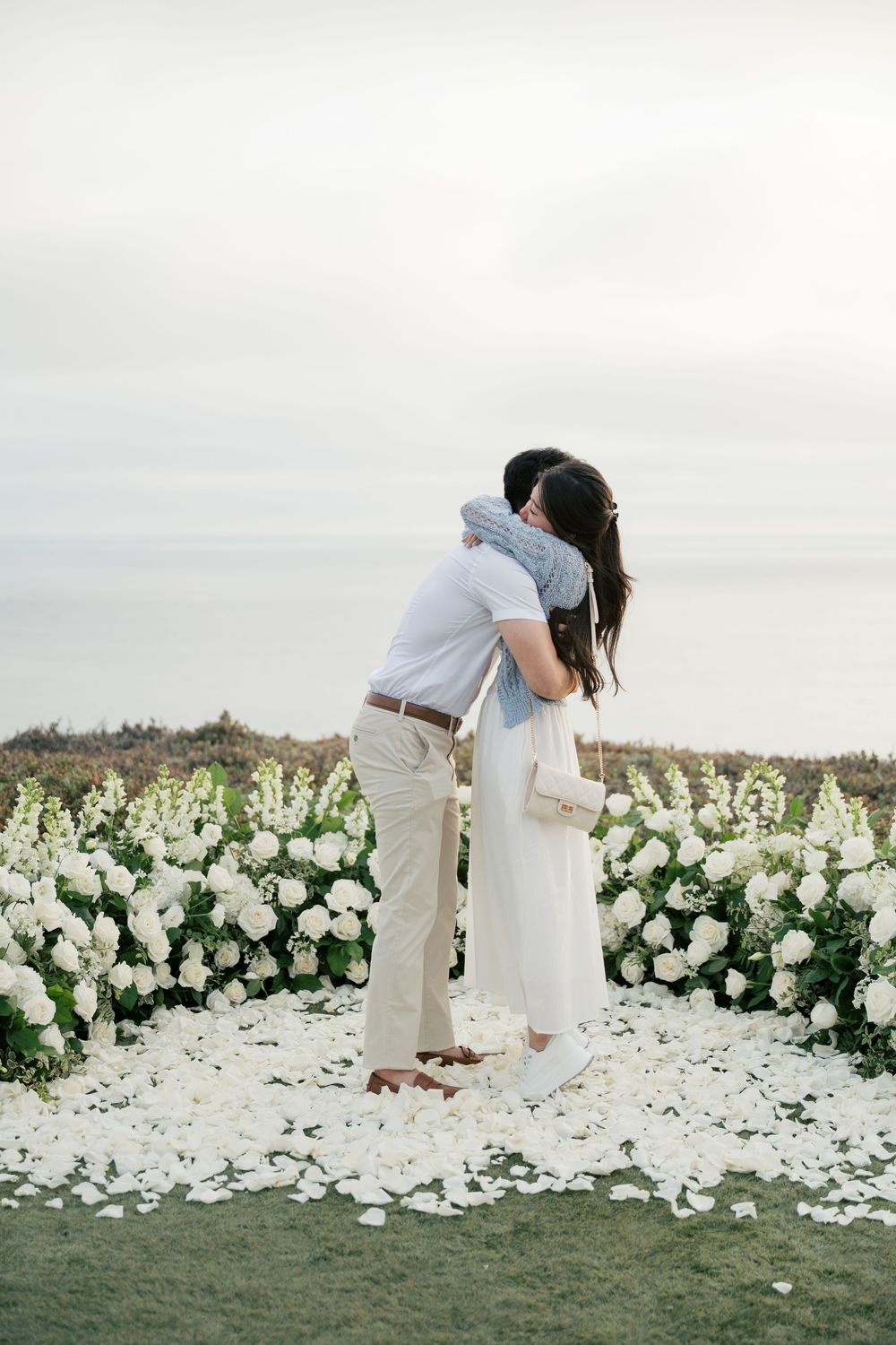 A couple embraces in front of a stunning display of white flowers on a grassy cliff overlooking the ocean.