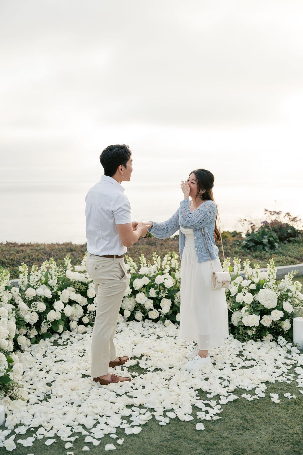 A romantic outdoor couple proposal scene with white floral decorations on grass against an ethereal background.