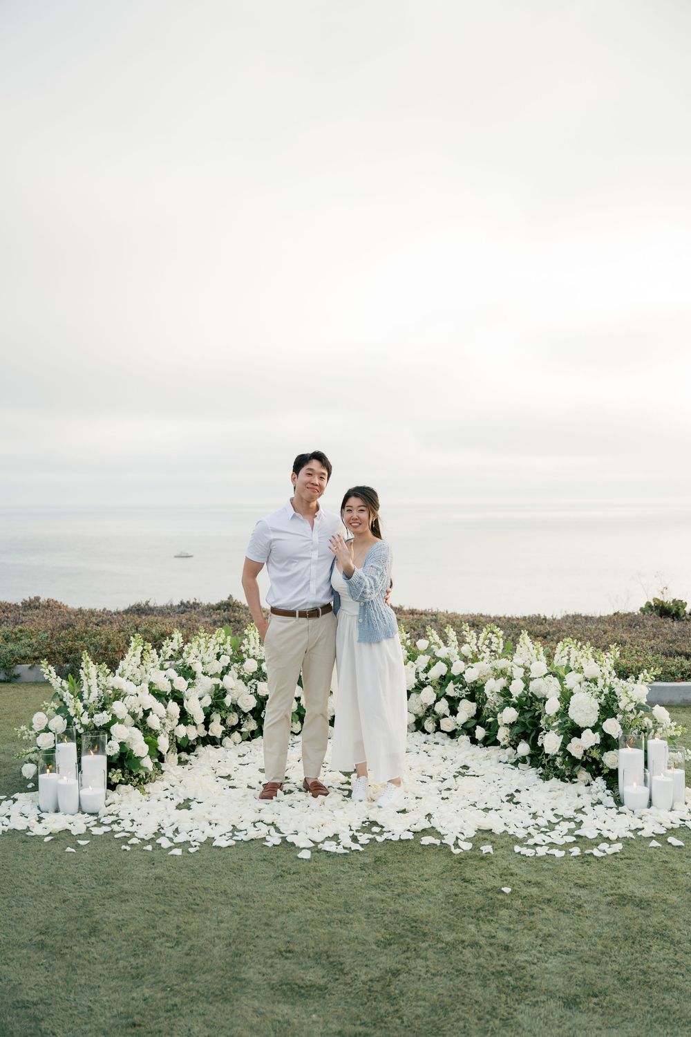 A couple stands together on a grassy coastline surrounded by elegant white floral arrangements at sunset.