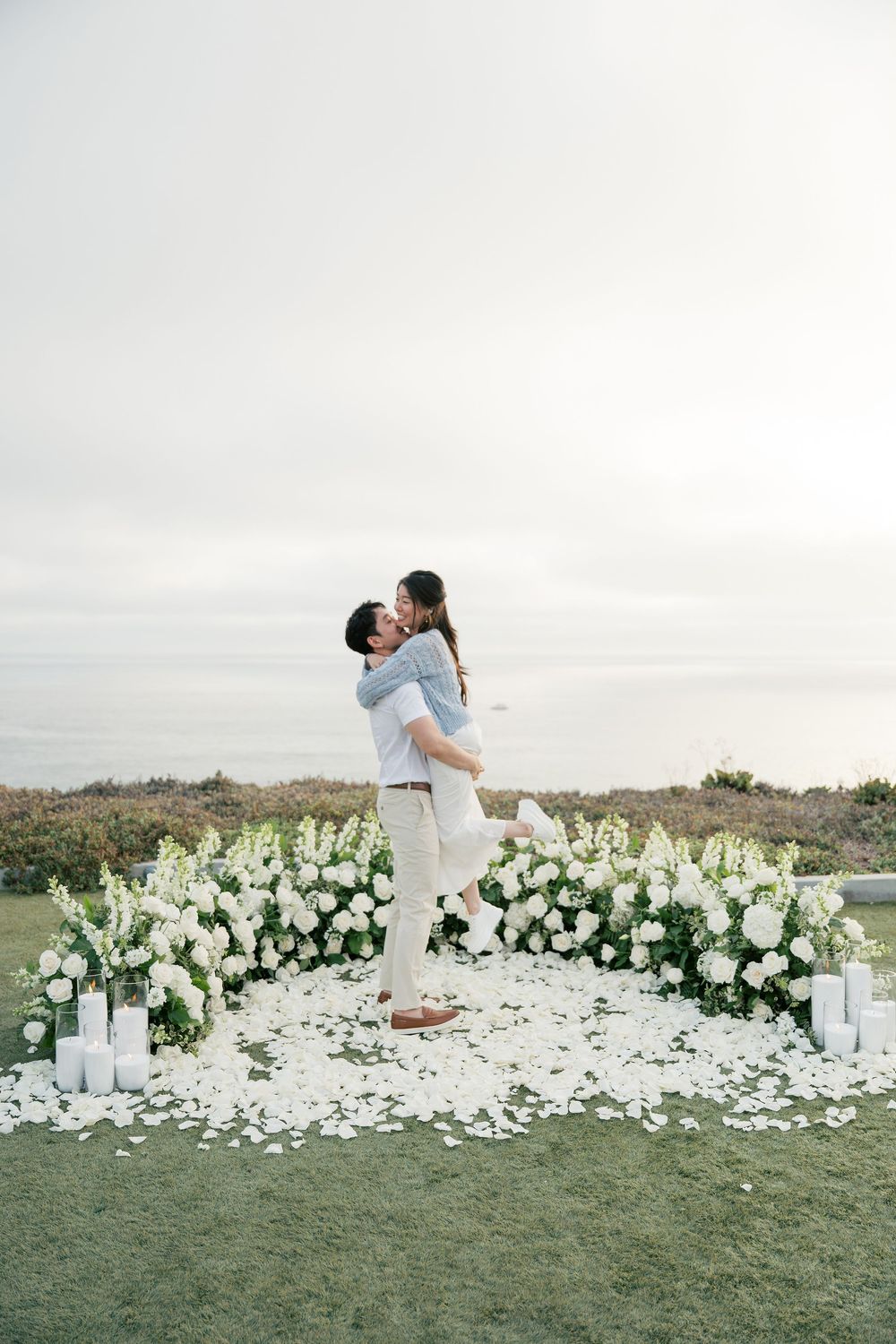 A romantic couple embraces outdoors surrounded by elegant white floral arrangements and candles at sunset.