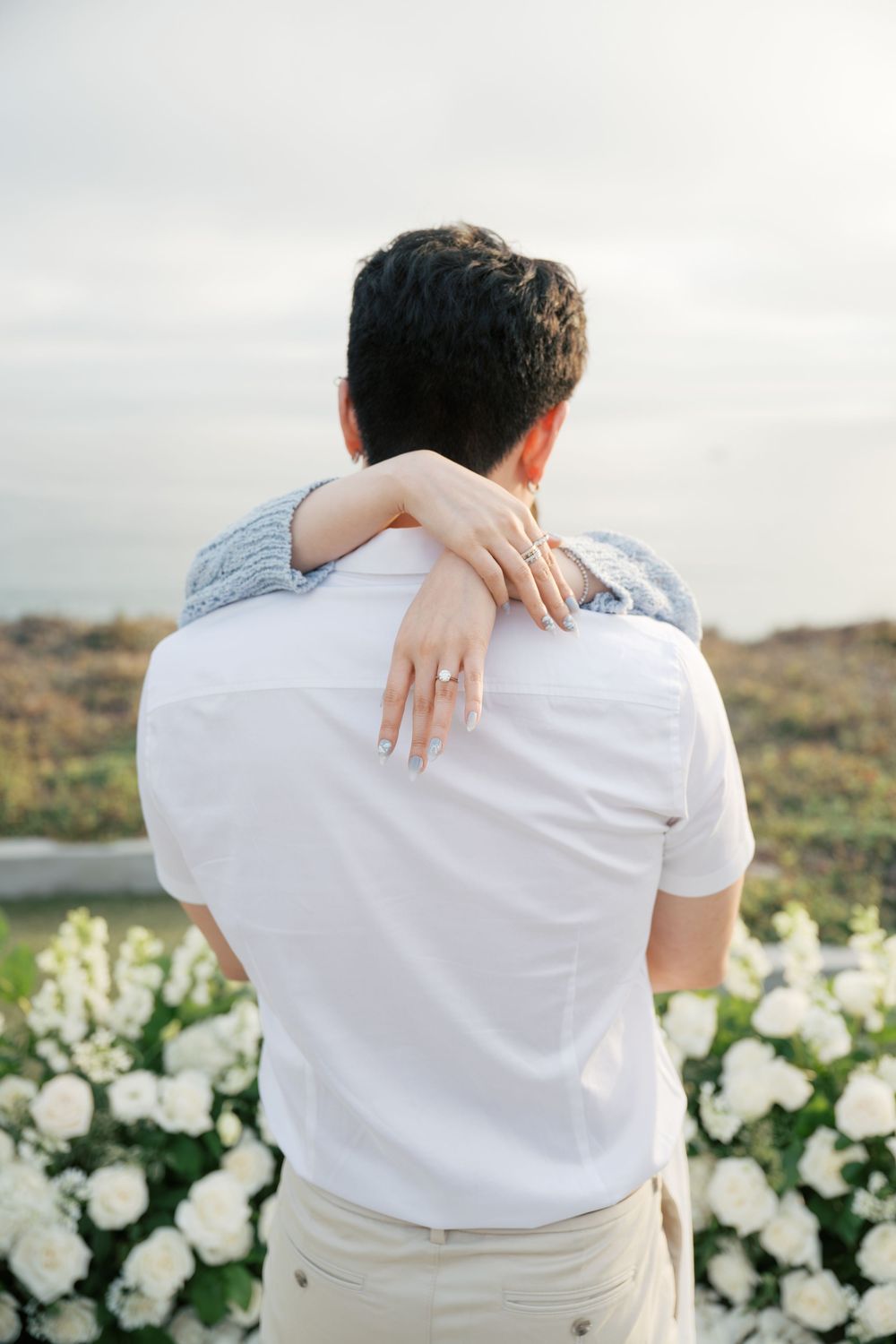 A couple in white embraces while sitting on an outdoor bench with white floral decorations behind them.
