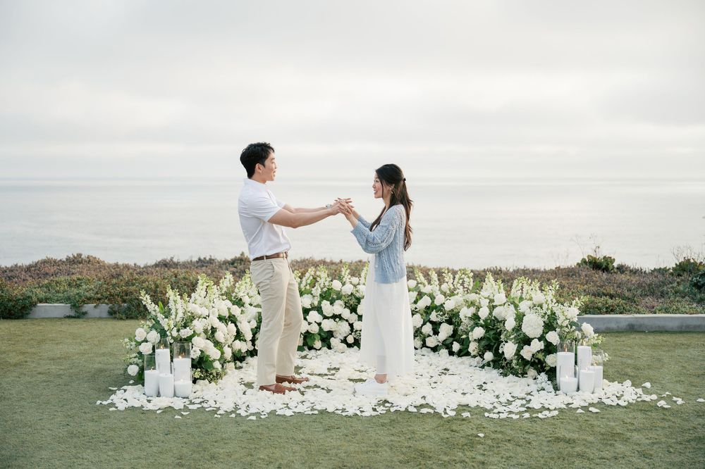A couple in white outfits poses for a romantic photoshoot in a landscape decorated with white floral arrangements.