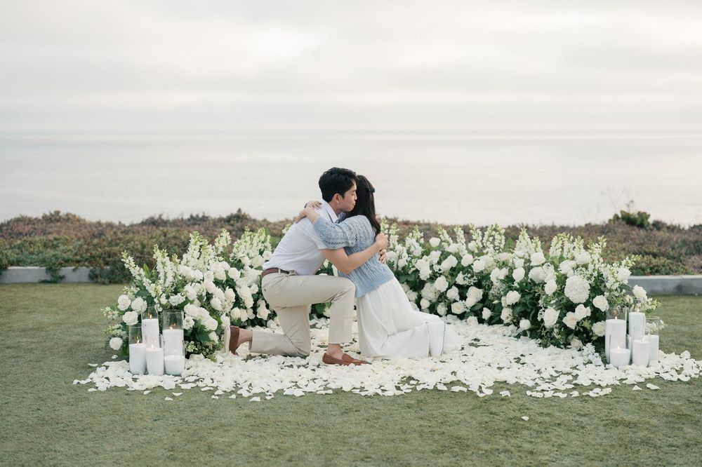A romantic outdoor wedding scene with a crescent shaped white floral installation and candles on a grassy hilltop at sunset.