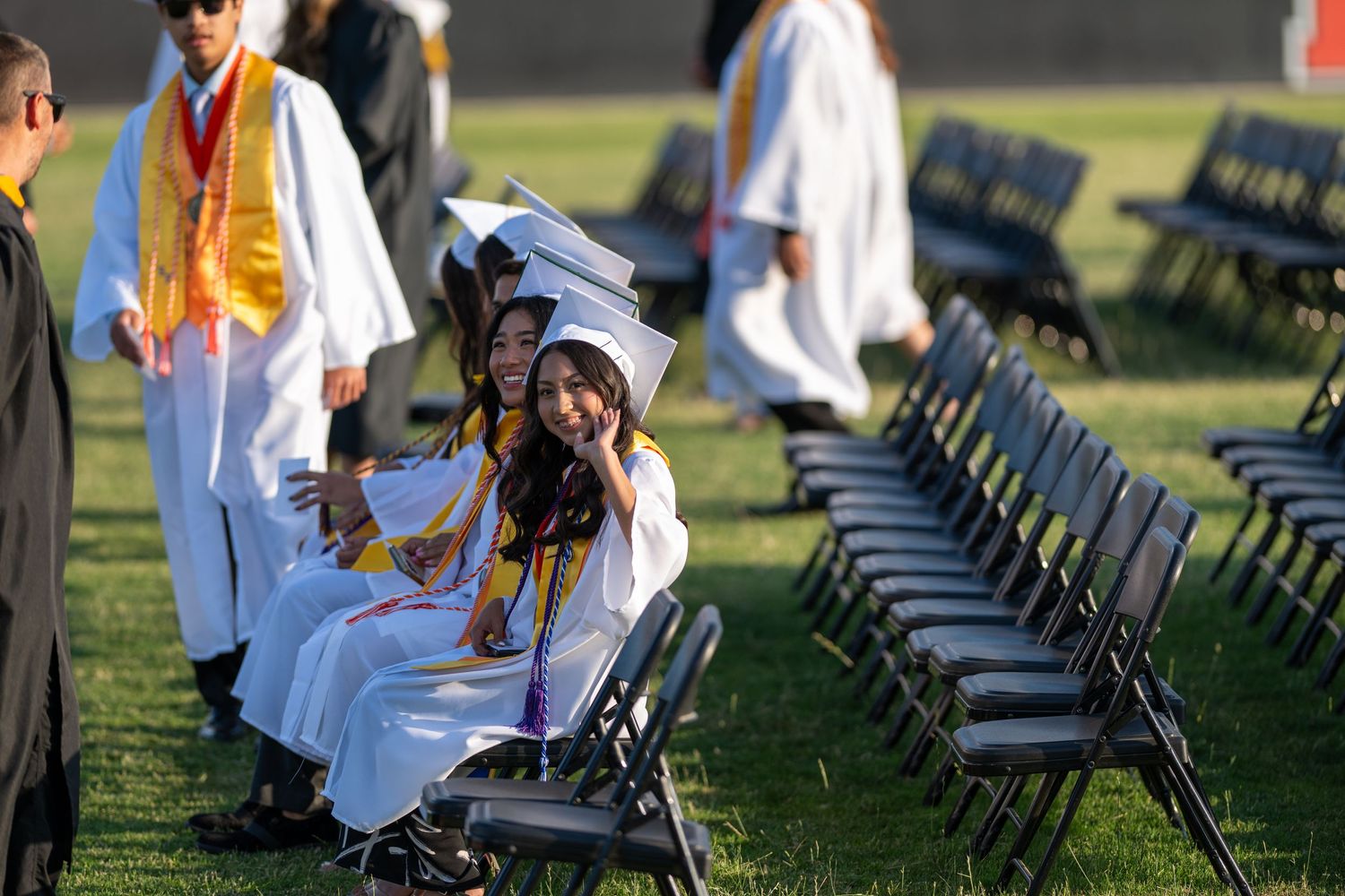 Graduates sitting in folding chairs during an outdoor commencement ceremony with bright sunlight in the background.