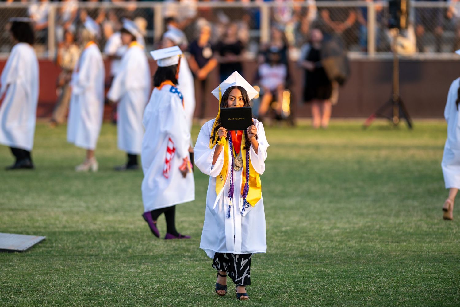 A line of graduates in white robes and caps walk across a green grass field during an outdoor graduation ceremony.