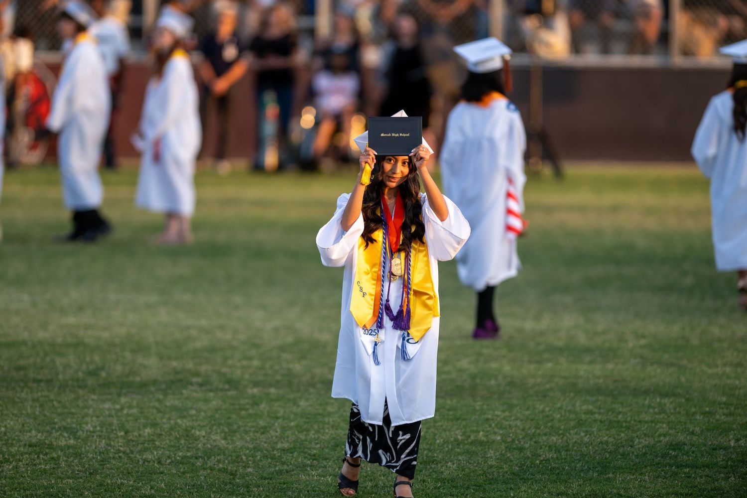 A graduate in a white gown and colorful stole takes photos during an outdoor commencement ceremony on a grassy field.